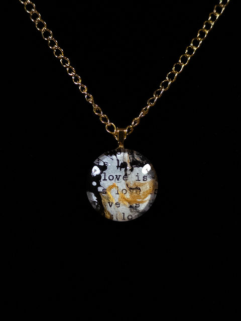 A necklace with a pendant made from a paper with typed words on a typewriter with paint swirls over the typed words, coved by a glass circle. Created by Emilyann Craighead