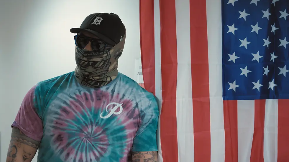 A masked ICE agent in a tie-dyed shirt leans against a wall bearing the American flag inside an immigration court.