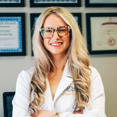 Woman with long blonde hair and glasses smiling, wearing a white coat in an office with framed certificates on the wall.