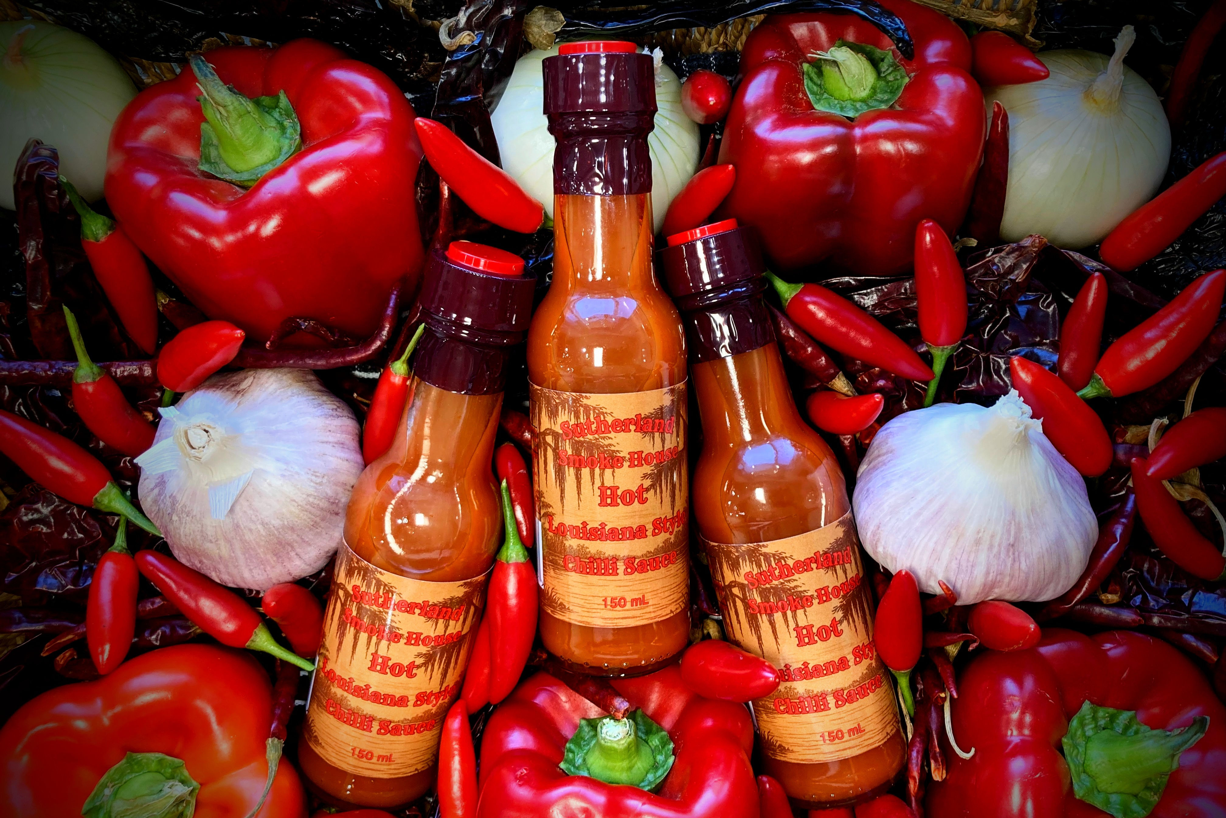 Three bottles of Sutherland Smoke House Louisiana-style hot chilli sauce surrounded by fresh and dry ingredients..
