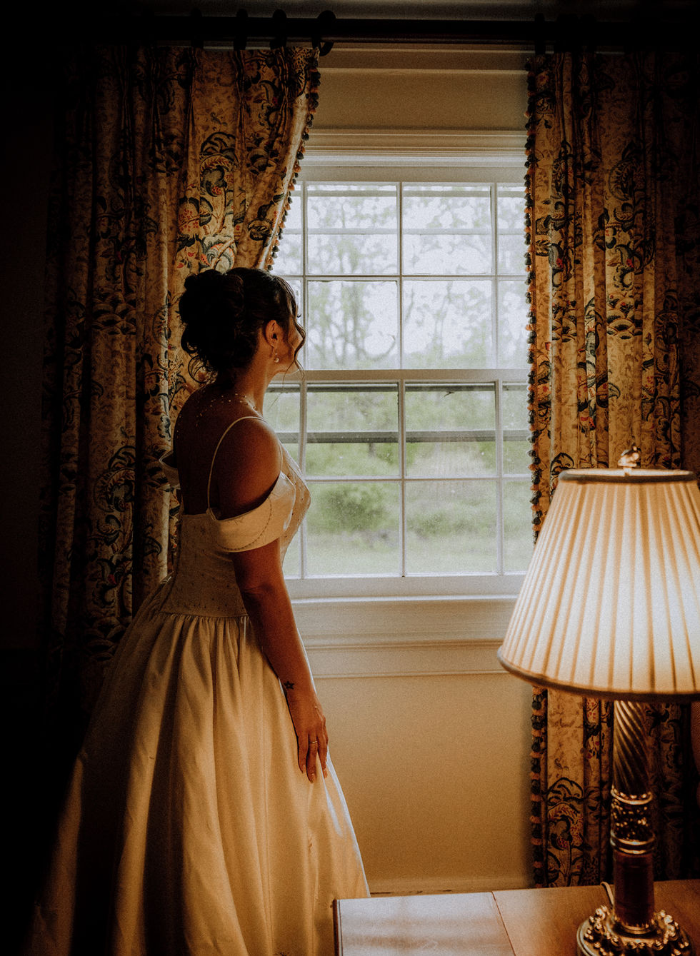 Bride looking out window beside lamp.