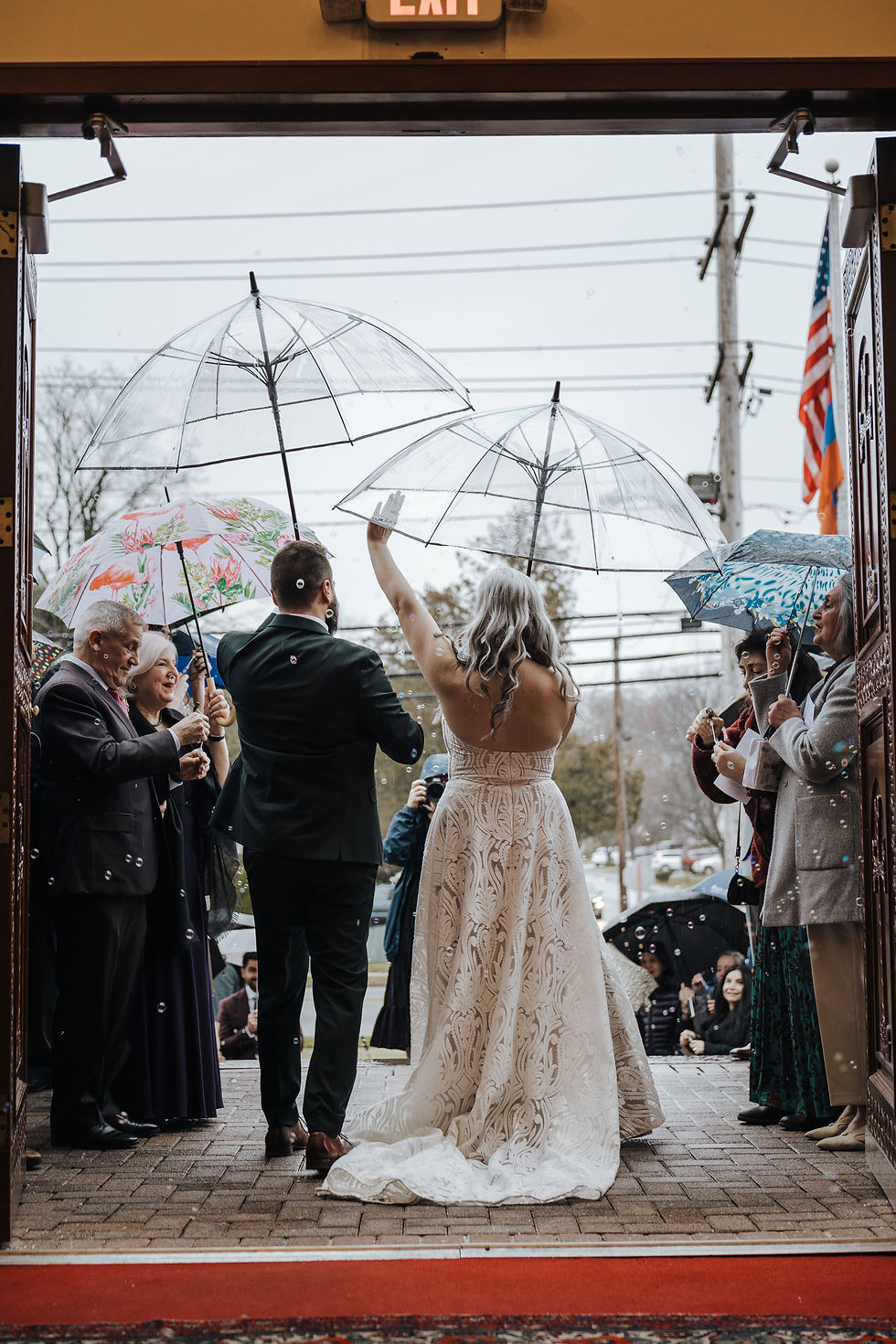 Bride and groom stand under clear umbrellas in a rainy church doorway as guests smile and blow bubbles.