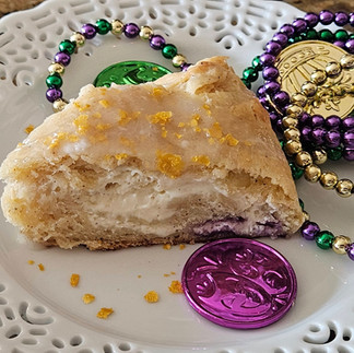 A slice of King Cake baked by Melissa Mitchell on a festive plate, revealing a cinnamon-cream cheese filling, surrounded by colorful beads and Mardi Gras doubloons.