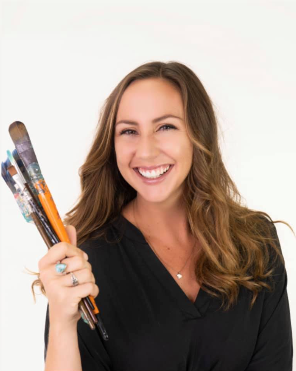 A smiling woman holding several paintbrushes, wearing a black blouse against a white background. This portrait captures artist Kaleigh Glover Ardoin, whose vibrant coastal paintings and murals celebrate color, creativity, and the beauty of Island life.