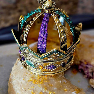 A homemade King Cake baked by Melissa Mitchell, topped with white glaze and sparkling gold sugar, adorned with a jeweled Mardi Gras crown in purple, green, and gold.