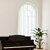 A photo capturing The piano is next to an ceiling height arched window draped with sheer c
