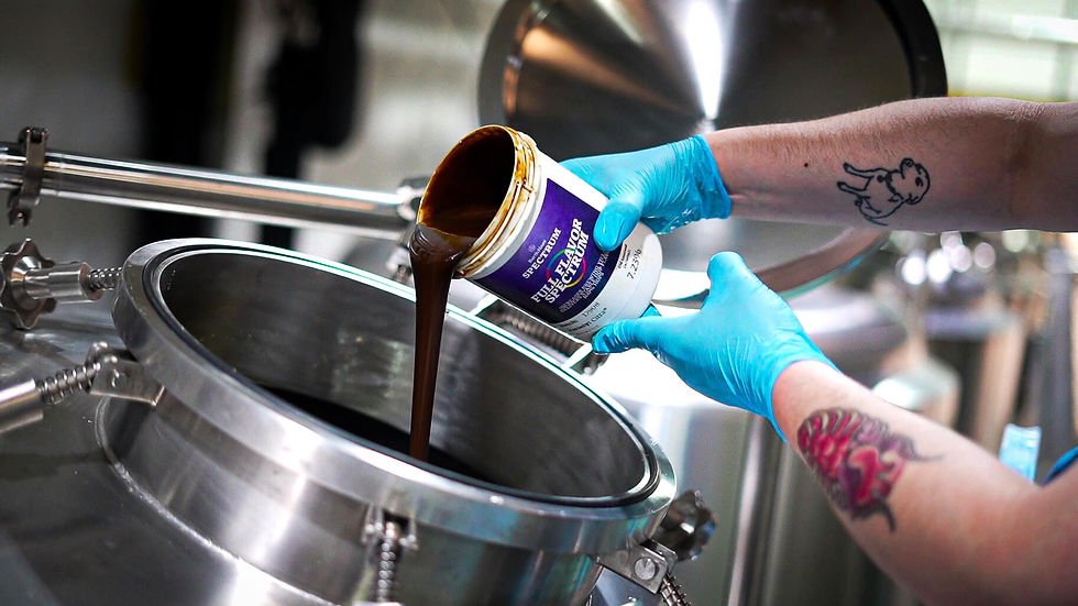 Gloved hands pour thick dark syrup from a Spectrum Flavor full-strength jar into a stainless steel tank in a brewery.