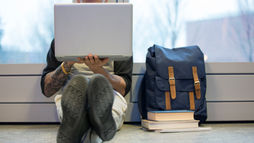 A youth sitting on the floor by a window, using a laptop with AI study tools, a blue backpack, books, and a notebook nearby. The mood is casual and focused, ideal for studying.