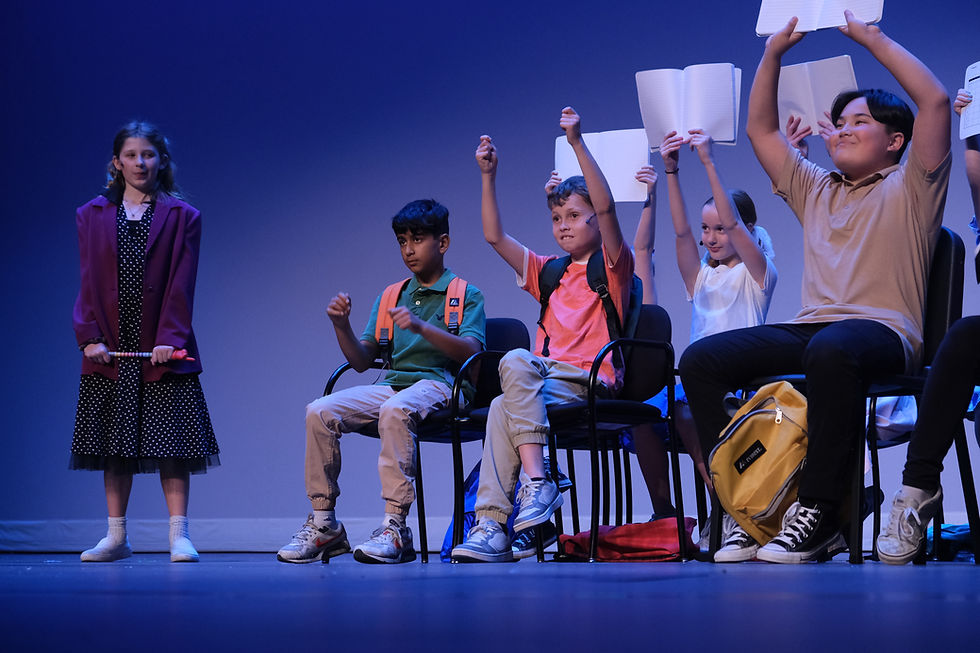 From 2024 Junie B. Jones, a teacher stands next to a class of sitting kids who are holding notebooks up over their heads