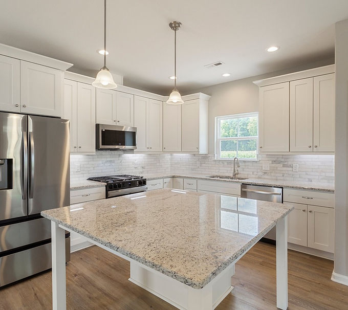 Kitchen for Pelaiah Model, white cabinets, brown wood lvp, white granite counter tops, kitchen island, two hanging light fixtures above island. Microwave above stove in wall, Stainless Steel Fridge left of stove and microwave,