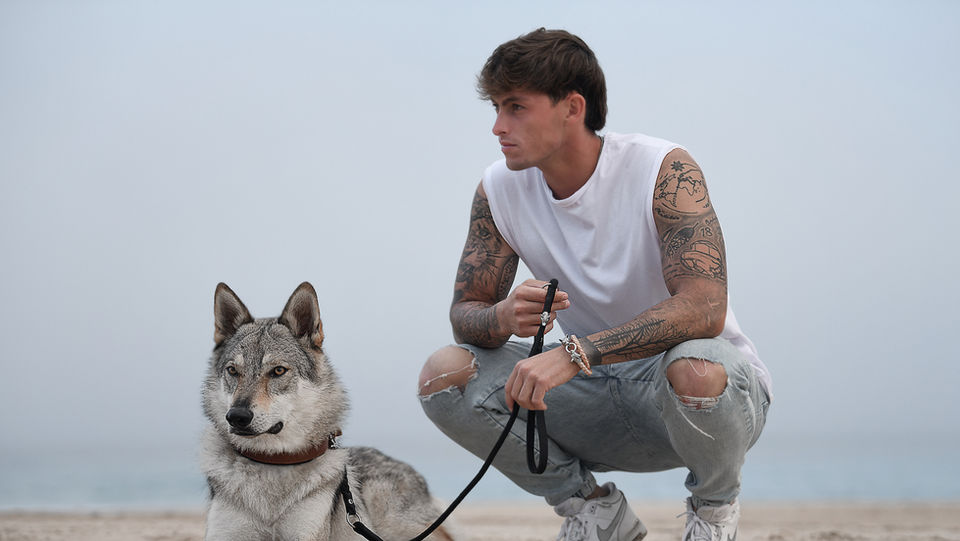 Fotografie eines hockenden Mannes mit einem angeleinten, liegenden Husky am Strand.