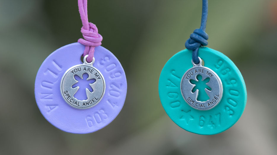 Alt-Text: Zwei bunte Hundemarken mit Gravur von KL Dog Accessories hängen nebeneinander vor unscharfem Hintergrund. Die linke Marke ist lila mit rosa Band und trägt den Namen ILUNA, die rechte ist türkis mit blauem Band und zeigt den Namen JAGO. Beide Marken sind mit einem silbernen Anhänger verziert, auf dem "YOU ARE MY SPECIAL ANGEL" steht.