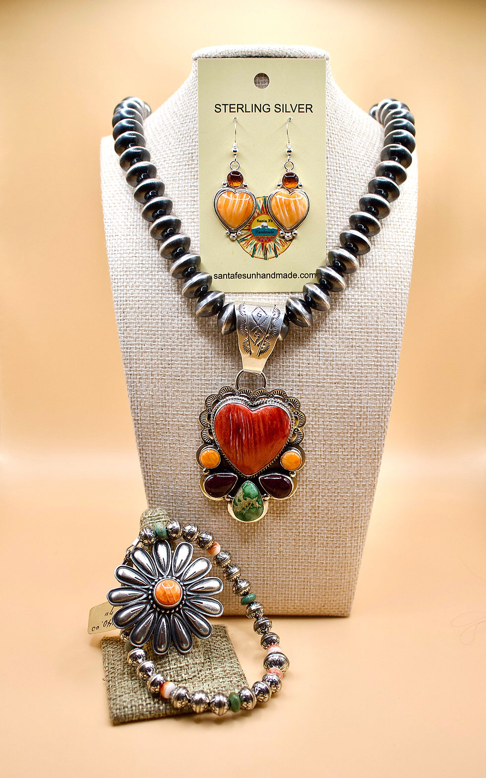 Sterling silver jewelry set on display: heart-shaped earrings, pendant with red heart, floral bracelet. Warm beige background.
