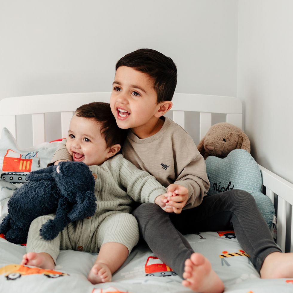 Two brothers, toddler and baby, sat on boys bed together, holding hands and smiling 