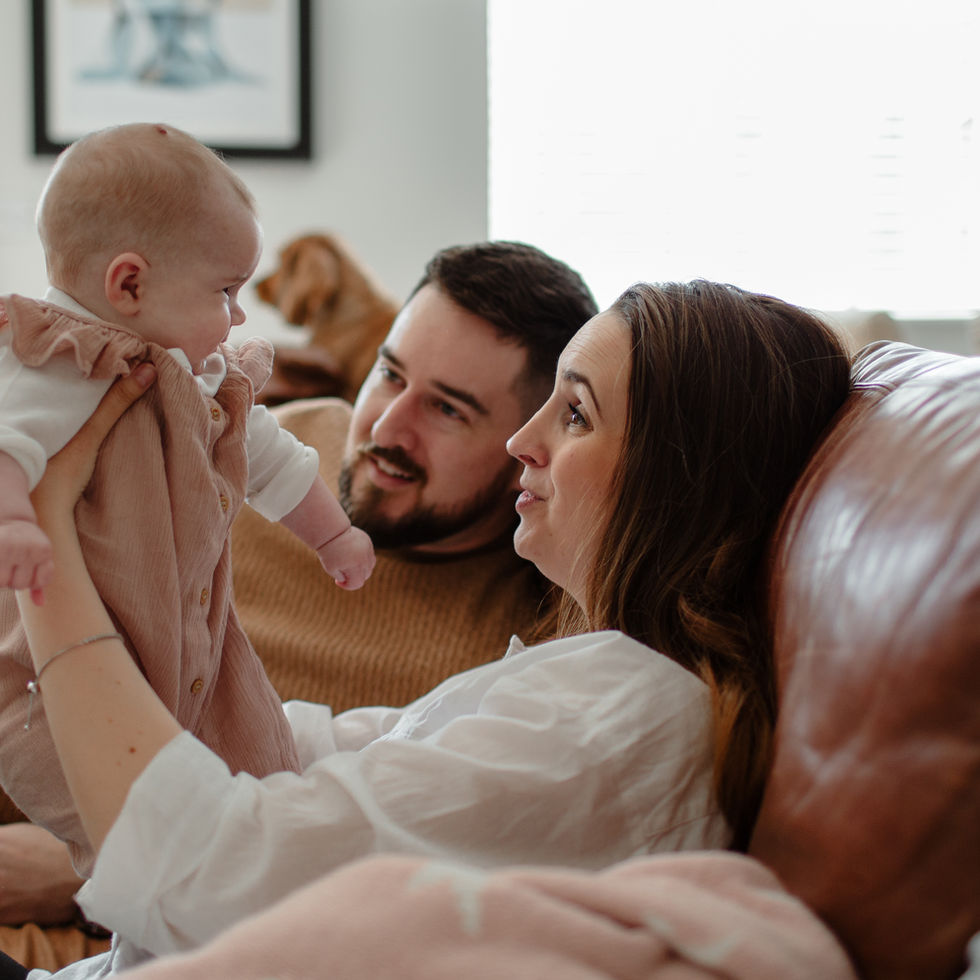 Mother and Father sitting on the sofa holding up 6 month old baby in dungarees.