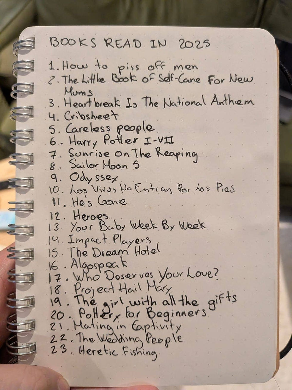 A full-page view of a handwritten list in a spiral notebook titled "BOOKS READ IN 2025." The list contains 23 numbered titles including "Project Hail Mary," "Mating in Captivity," and "Heretic Fishing." A person's thumb is visible on the left edge, holding the notebook open.