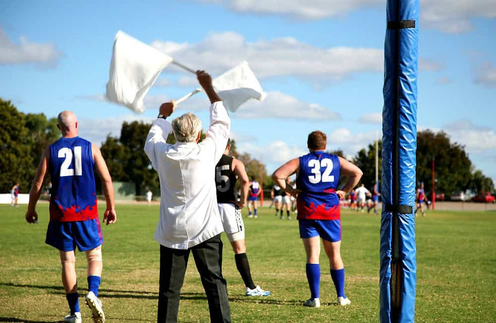 Australian Rules football game in Melbourne with goal umpire signalling a score, players wearing blue and red jerseys on a sunny day.