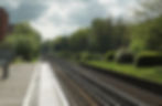 Train tracks at Walton-On-Thames train station stretch into the distance beside a lush green forest and empty platform. Overcast sky adds a moody atmosphere.