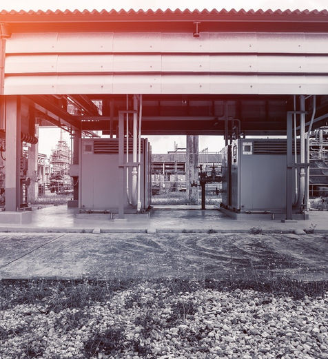 Exterior, low-angle, black and white photograph of two industrial packaged compressor or pump stations housed under a metal-roofed shelter in an outdoor plant setting. The equipment enclosures are visible beneath the roof, and the foreground shows a gravel and dirt patch, with complex industrial piping visible in the background. A strong sun flare adds an orange tint to the roofline.