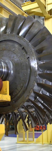 Close-up of a massive industrial gas turbine rotor lying horizontally in a brightly lit factory. The rotor is cylindrical, featuring hundreds of precisely angled, shiny metal turbine blades arranged in multiple stages. The large metal shaft is supported by heavy-duty yellow industrial stands and equipment, indicating it is undergoing assembly.