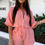 Thumbnail: Woman wearing a coral belted cotton gauze playsuit with white sandals and a white mini handbag, standing on a cobblestone str