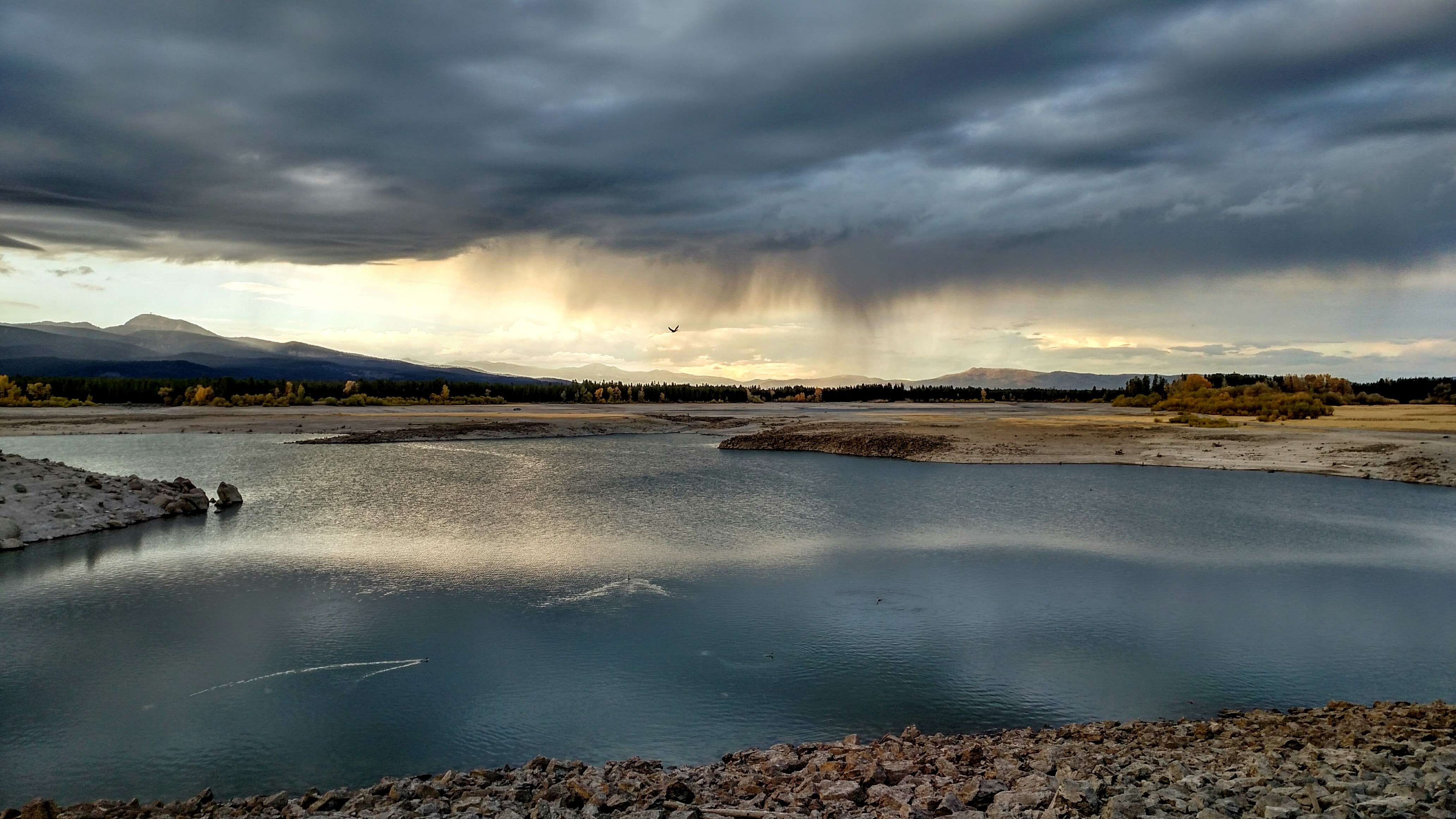 Island Park Reservoir with low water levels and exposed sediment that is typically underwater. The sky is stormy and blocking the afternoon sun. The first wisps of rain are starting to fall.