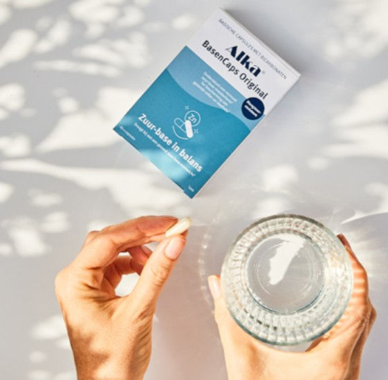 Thumbnail: Een hand houdt een capsule vast naast een glas water, met een verpakking van Alka BasenCaps op een lichte achtergrond.