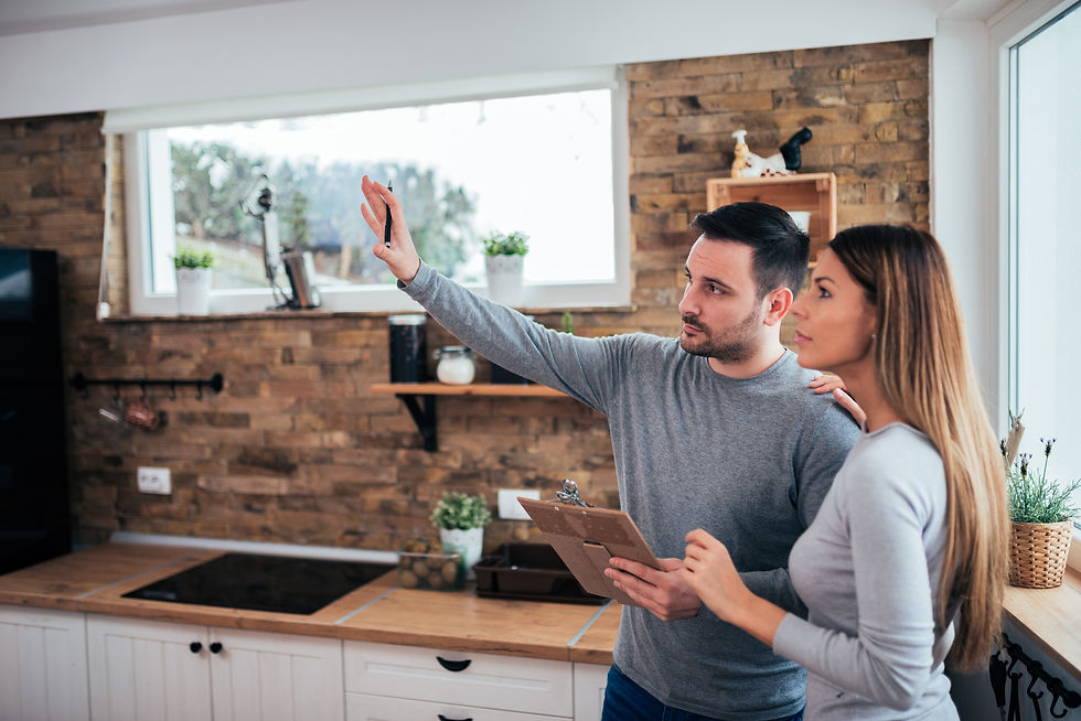 Two people in a modern kitchen, discussing plans. One holds a clipboard, gesturing toward a window. Brick wall and white cabinets visible.