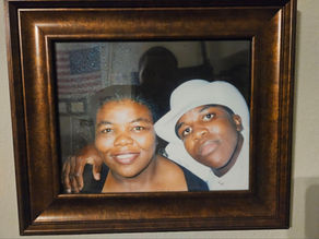 Framed photo of two people smiling, one wearing a white hat. An American flag is faintly visible in the background, brown wooden frame.