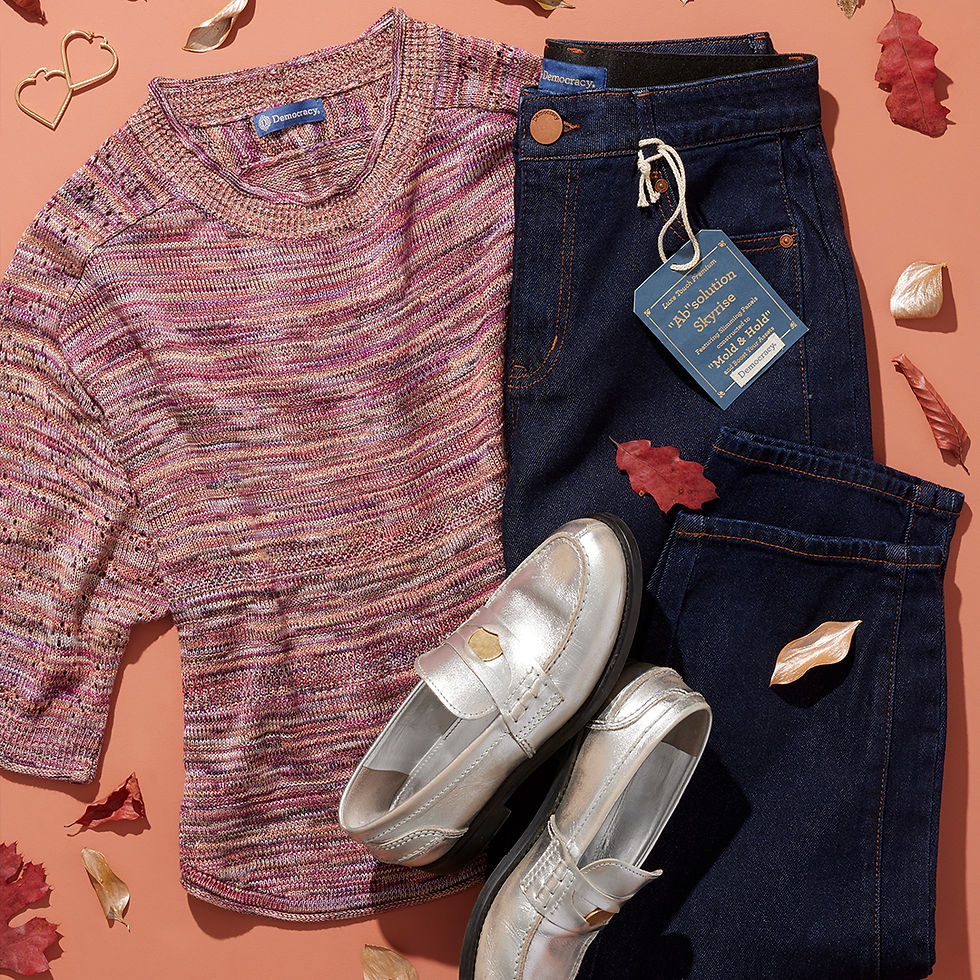 Flatlay of a pink knit sweater, dark blue Democracy jeans, and metallic loafers styled with fall leaves on a peach background.