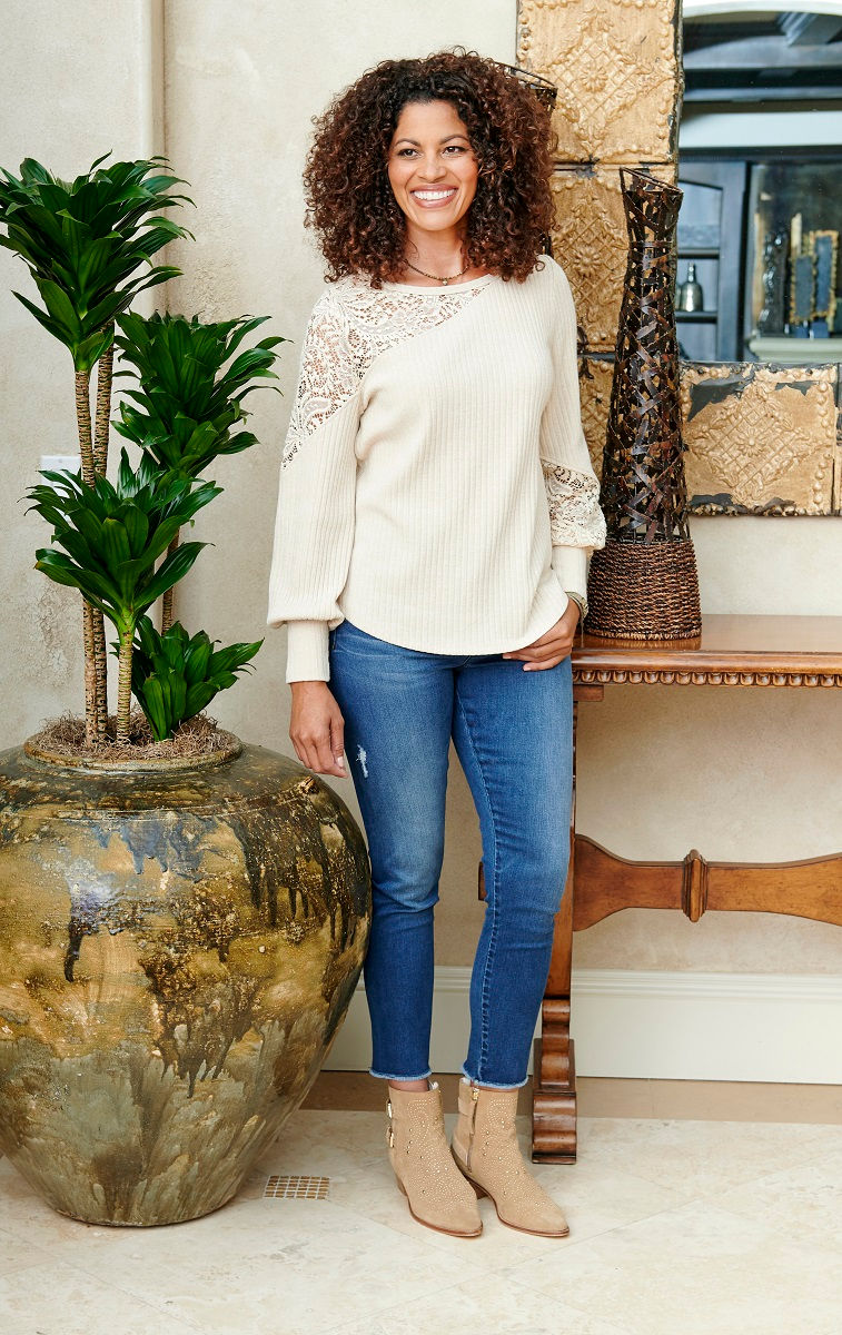Woman wearing a cream knit sweater with lace detail on the sleeves and blue jeans, standing indoors beside a decorative plant and console table.