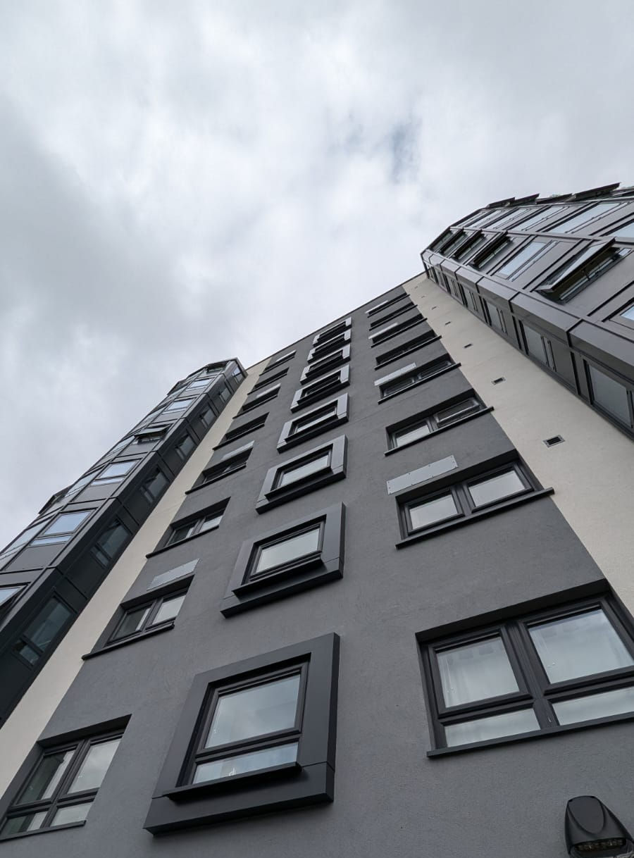 High-rise residential building facade with dark grey render and projecting aluminium window sills, viewed from below.