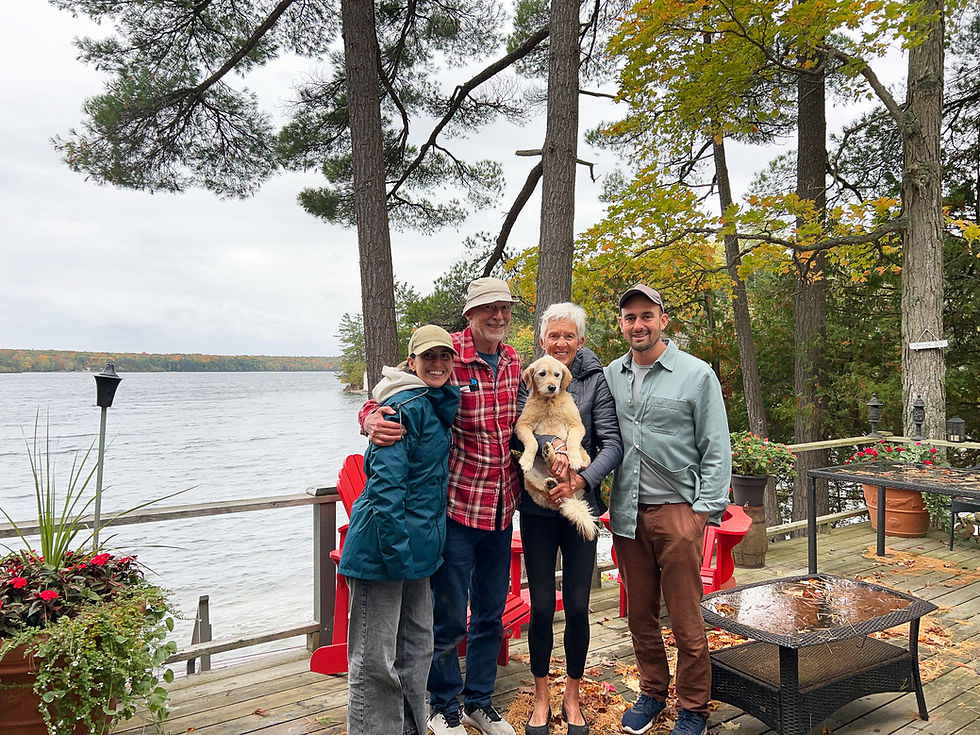 Family with dog stands smiling on a deck by a lake surrounded by autumn trees. Red chairs and a table are in the background.