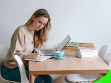 Woman working at a desk with a laptop, writing in a notebook, and enjoying a cup of coffee.