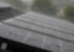 Rain splashes across dark solar panels on a rooftop, with blurred trees and houses in the background.