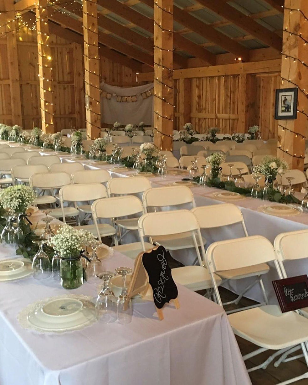 Wedding Reception ready for Guests at Grand Junction Barn Venue. White tables cloths with greenery and flowers on the tables and twinkling lights wrapped on wooden post in barn.