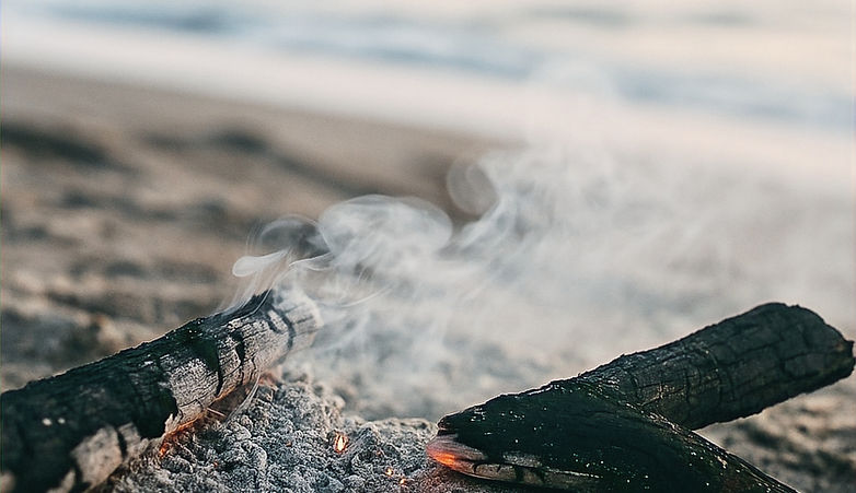 Close-up of charred wood and smoke rising from a smouldering fire on the sand, with a soft-focus ocean in the background