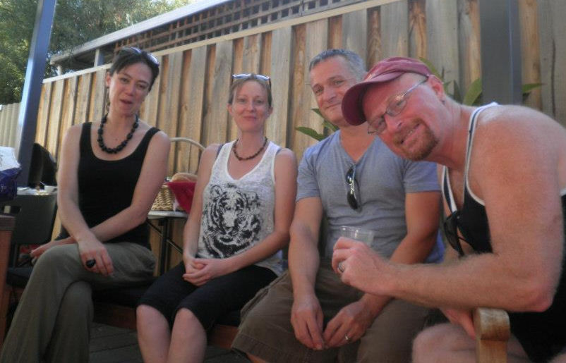 Four people sit on a patio bench, smiling, with a wooden fence behind. One person wears a red cap, another a tiger print shirt. Casual setting.