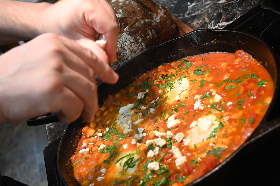 Hands sprinkle cheese over shakshuka in a black skillet. Tomato sauce and herbs visible. A loaf of bread is in the background. Cozy setting.