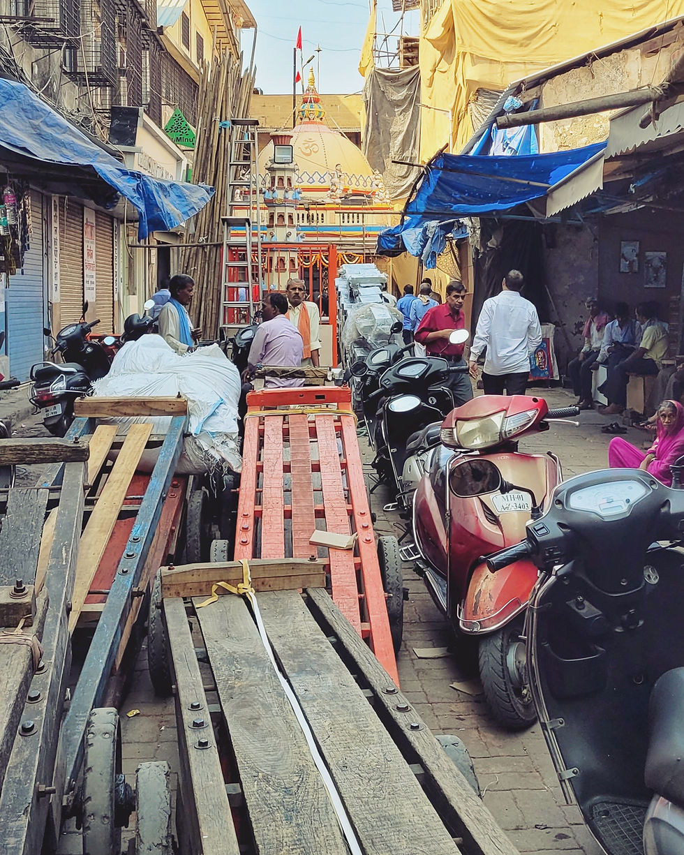 How do our technologies shape us and our lives?
That’s a question I really started asking myself during my trip to Mumbai, India, and ever since. It was the farthest I’d ever been from home and my first time in Asia. One of my favorite things to photograph there was the way recognizable technologies appear in unrecognizable surroundings, areas, and locations—or sometimes the other way around.
I was struck by the level of artisan lifestyle, which says a lot coming from the Caribbean. At the same time, so much also felt familiar. Many places I’ve lived in and traveled to in the Caribbean have Indian populations and influences, so parts of Mumbai resonated with me in unexpected ways.
Next, I’ll highlight some of the photos from my gallery Bom Baim. Alongside them, I invite you to reflect on a critical question: Do standardized technologies shape, help, or add to societies in unique ways? Or do they also risk unifying—and perhaps negatively uniforming—our world, making it feel monotonous?
Two corners away from my hostel—which was in an Islamic neighborhood in the slums, with no English speakers, and where I had slept alongside eleven other men in bunk beds—I walked past this butcher shop at 6 am. If you look closely, what might seem bizarre to some are the live goats standing right next to the slaughtered and displayed ones. What chaos—and what to imagine awaits them! Yet, on the tiled wall behind, there’s a QR code for Paytm, allowing people to buy meat digitally with their phones. It's a juxtaposition and provides a glimpse of how India employs technologies.
A few streets away, I spotted a strikingly modern high-rise—sleek, tall, with flawless finishing. From far, it looked like precision architecture and engineering. Then I noticed the scaffolding: not steel or aluminum, but bamboo. The tall, wiggly stalks wrapped around the building all the way to the top, supporting construction as it was being finished. It was a fascinating contrast: cutting-edge design held up by an ancient, natural material. An inversion of what we usually associate with technology—where bamboo, flexible and organic, replaces the heavy, industrial structures we expect.
Just a little further, at the end of a narrow, bustling lane, I came across a small temple. It offered another striking example of how technology is processed in Indian society. On one side, human-driven carts were parked—simple, manual, and labor-intensive. Right beside them, scooters, sleek and motorized. What caught my eye was the unexpected harmony: despite the vast difference in power and speed, the carts and scooters shared a visual language—they were both painted in shades of red and black, creating an almost poetic symmetry amid the disparity. It was a vivid reminder that technology doesn’t just exist in isolation; it interacts with society.