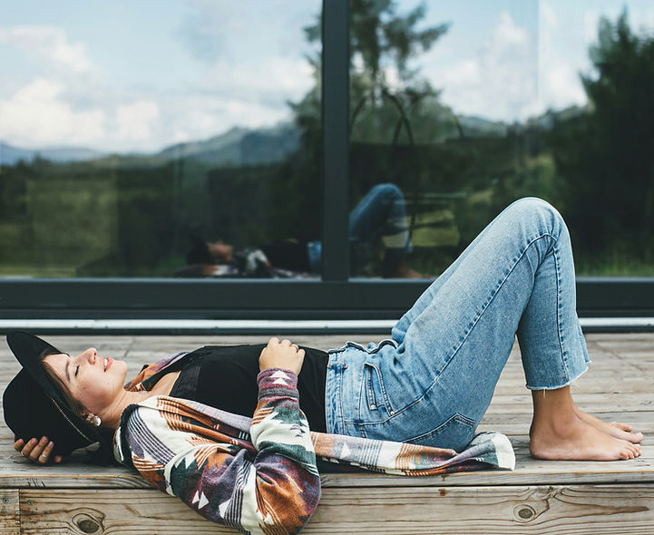 Casual image of a woman lying back on a deck, hat covering her face, against a mountain backdrop. Text describes the lived reality of therapy: crawling in, mumbling through tears, or making a dark joke.