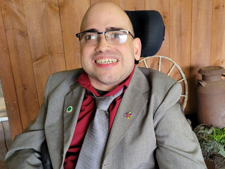 Kevin is smiling, wearing glasses, a grey suit and tie and is in his wheelchair.