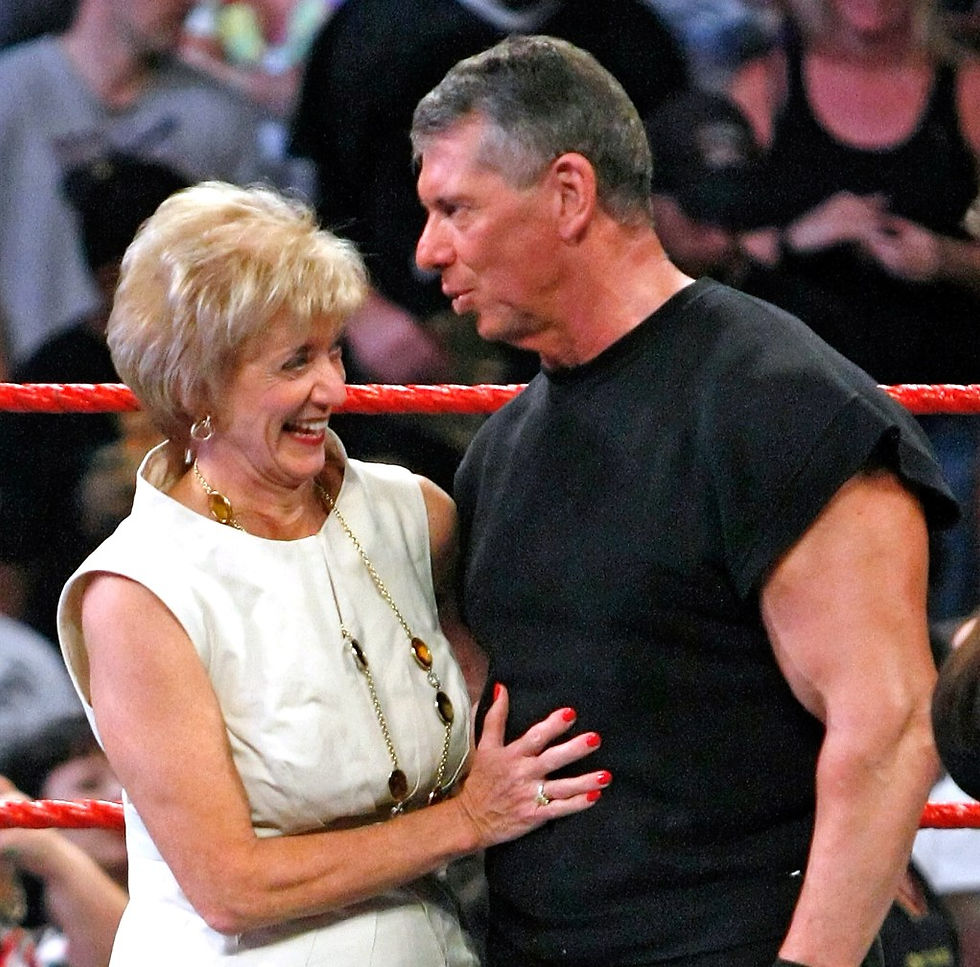 A blonde older woman with beach blond hair is smiling, wearing big jewelry and a sleeveless white dress, standing next to a tall, older grey haired man wearing a black muscle shirt.