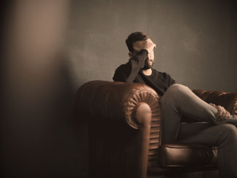 A man with dark hair sitting on a leather lounge with his head hung in his hand looking worried and moody