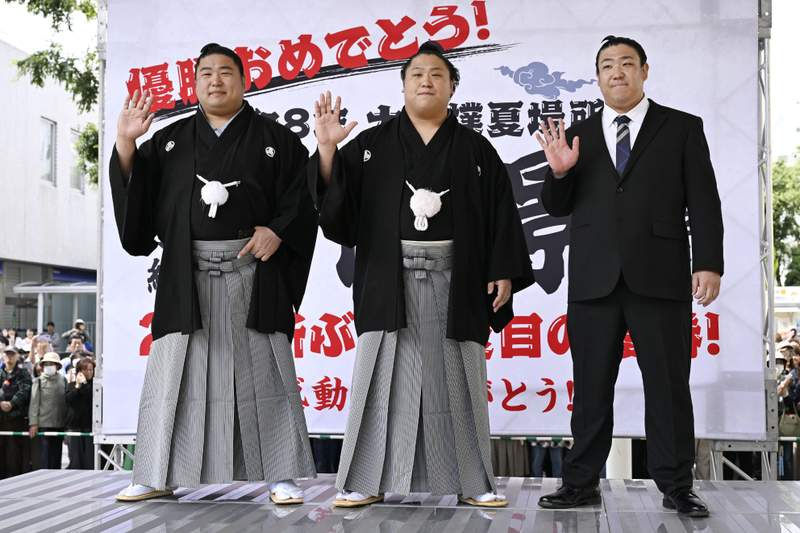 Deux lutteurs de sumo et un homme en costume saluent sur scène, devant une grande bannière japonaise festive.