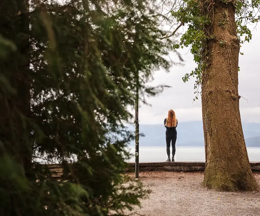 Frau blickt zwischen Bäumen auf einen See und Berge.