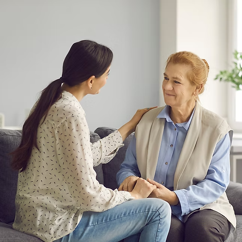 two women seated on grey sofa—younger woman comforting older woman with hand on shoulder