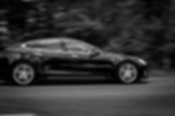 A black Tesla electric car with new tires driving fast on a road in front of a blurry background