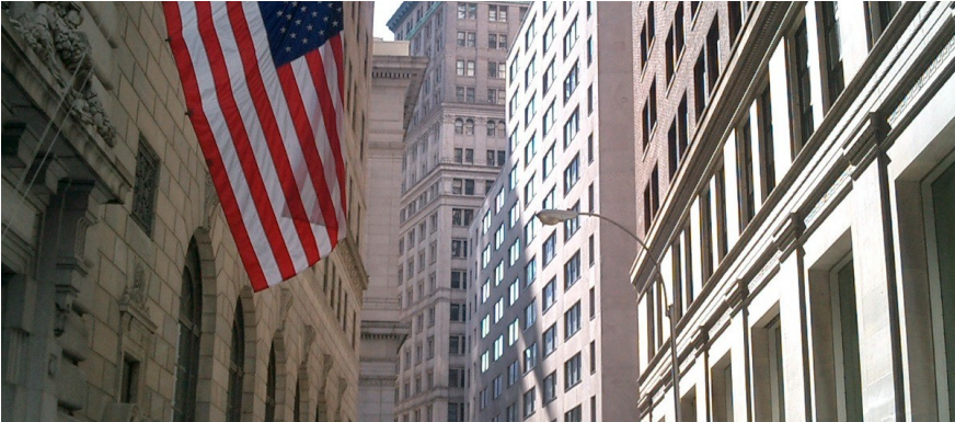 American flag hangs between tall downtown buildings on a sunny city street. Wall Street