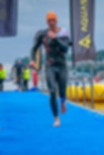 Athlete in wetsuit and orange cap runs on blue mat. Background shows water, safety personnel, and flags reading "AQUASPHERE." Focused mood.